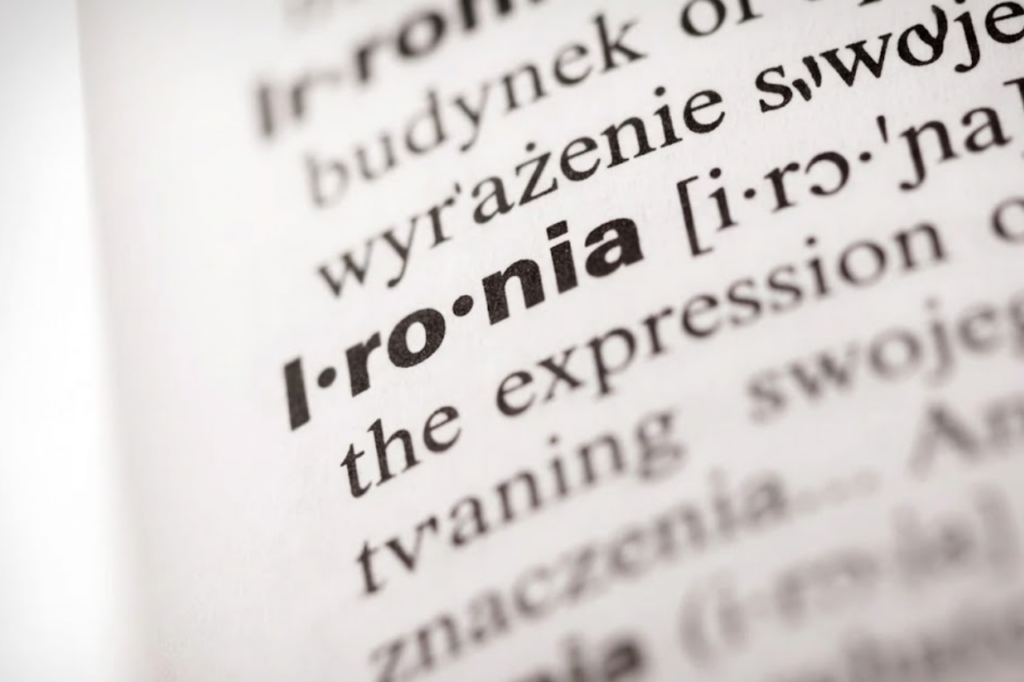 Zbliżenie na słownikową definicję słowa "ironia" wydrukowaną na białym papierze, z widoczną transkrypcją fonetyczną i fragmentem opisu znaczenia.