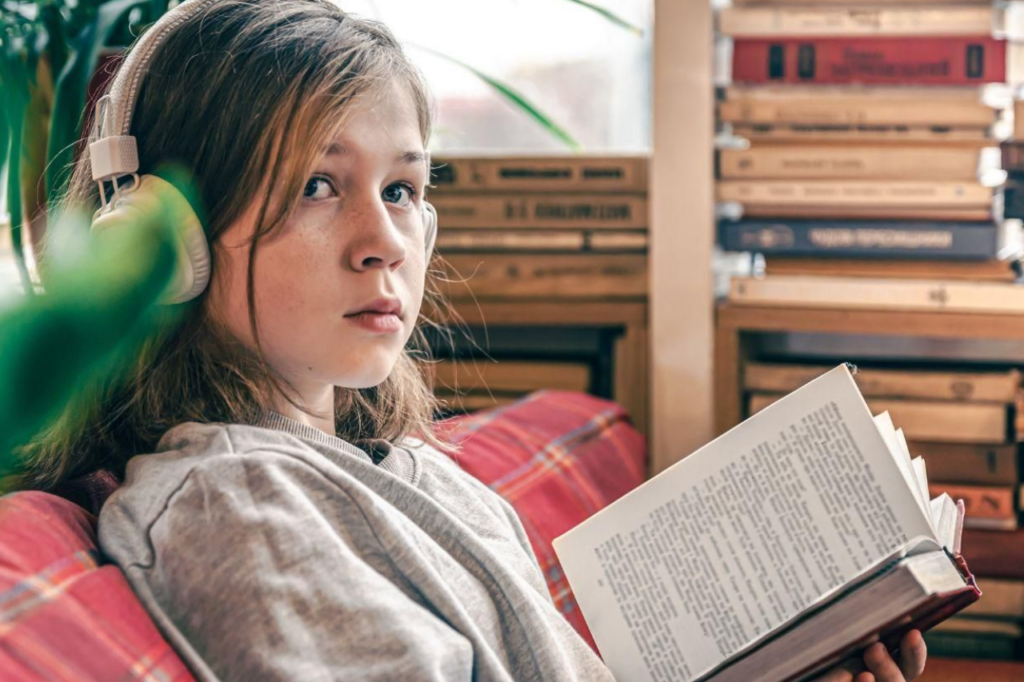 Młoda osoba w słuchawkach czytająca książkę na tle domowej biblioteczki, ilustrująca etap analizy postaci do wypracowania z języka polskiego.
