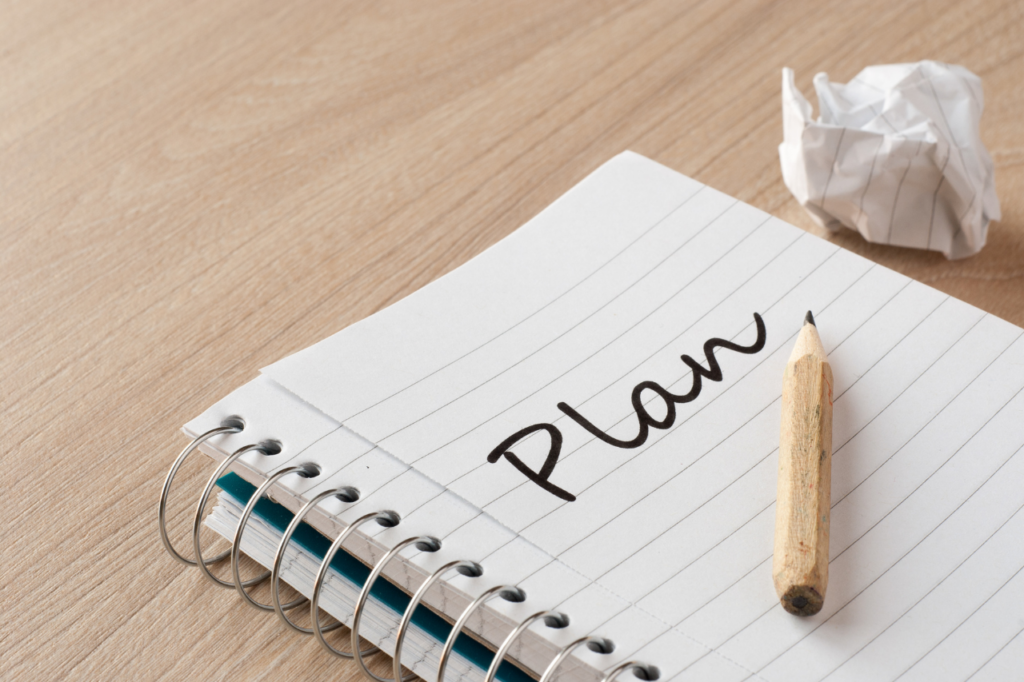 Notes na spirali z napisem "Plan" na pierwszej stronie, obok którego leży krótki ołówek i zgnieciona kartka papieru na drewnianym blacie.