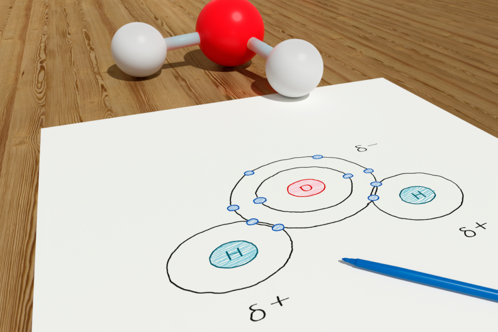 Trójwymiarowy model cząsteczki wody obok kartki papieru z odręcznym schematem struktury atomowej, wiązań chemicznych i polaryzacji, ilustrujący proces nauki podstaw chemii.