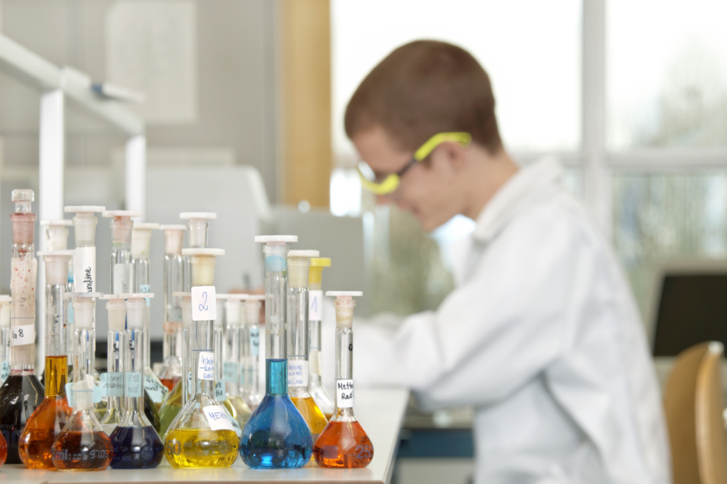 Kolorowe roztwory chemiczne w kolbach miarowych na blacie laboratoryjnym na tle pracującego ucznia w okularach ochronnych, obrazujące praktyczne metody rozpoznawania kwasów, zasad i soli.